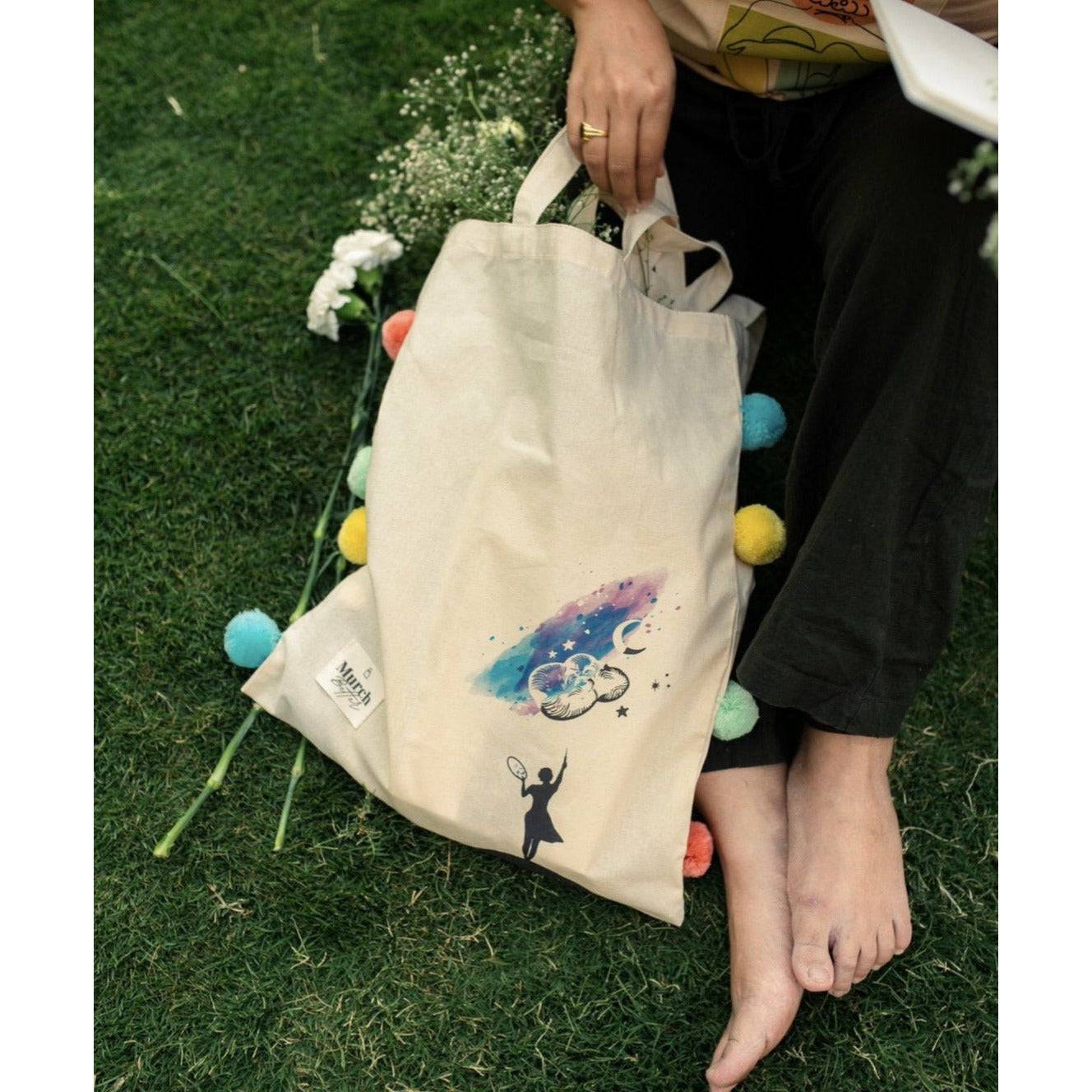 Person holding a tote bag with pom-poms and celestial artwork